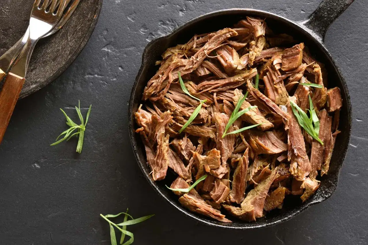 Soczyste pulled pork w żeliwnej patelni, gotowe do podania. Obok widelce i świeże zioła.