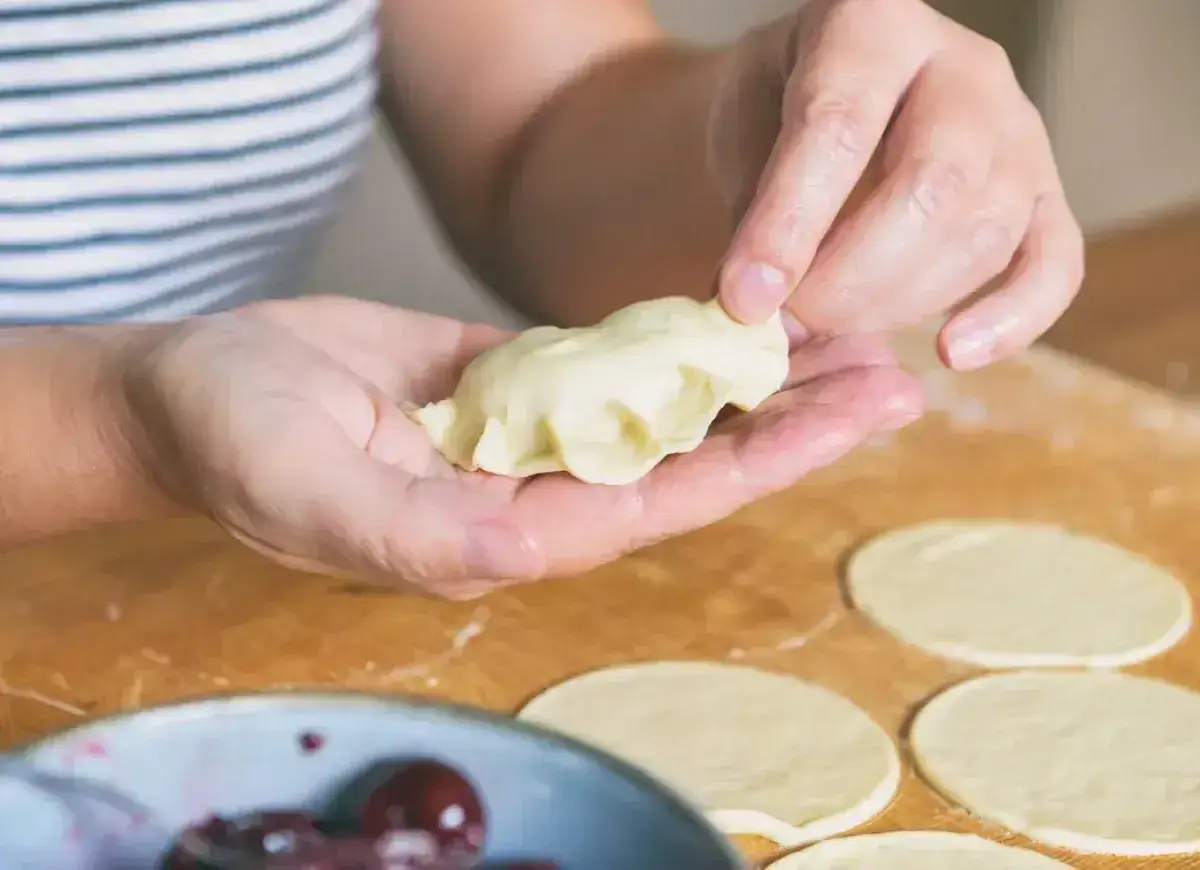 Dłonie formują pieroga, pokazując jak zrobić pierogi. Na blacie leżą okrągłe placki ciasta i miseczka z wiśniami.