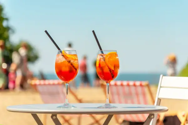 Dwa orzeźwiające aperol z tonikiem na stoliku plażowym, w tle morze i ludzie.