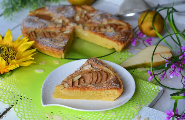 Pyszna tarta z gruszkami, posypana cukrem pudrem i płatkami migdałów, prezentuje się apetycznie na talerzyku. Obok cała tarta, gruszki i słonecznik.