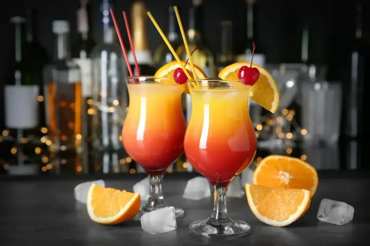 Dwa drinki z pomarańczą i wiśnią, przypominające Tequilę Sunrise. W tle bar z butelkami i światłami.